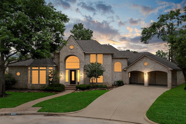 french country inspired facade featuring a front lawn, brick siding, a shingled roof, concrete driveway, and an attached garage