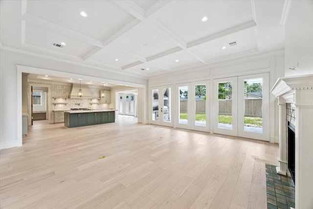 unfurnished living room featuring healthy amount of natural light, french doors, a fireplace with flush hearth, light wood-style flooring, and coffered ceiling