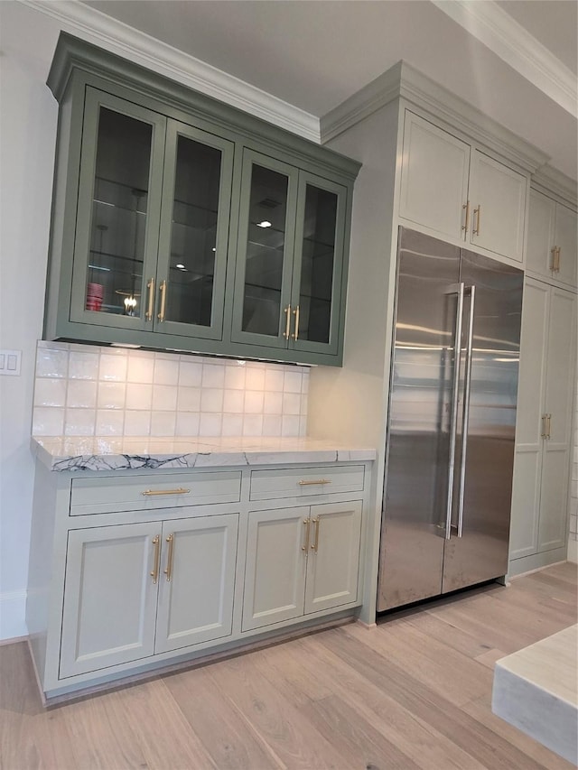 kitchen featuring stainless steel built in refrigerator, light wood finished floors, crown molding, backsplash, and glass insert cabinets