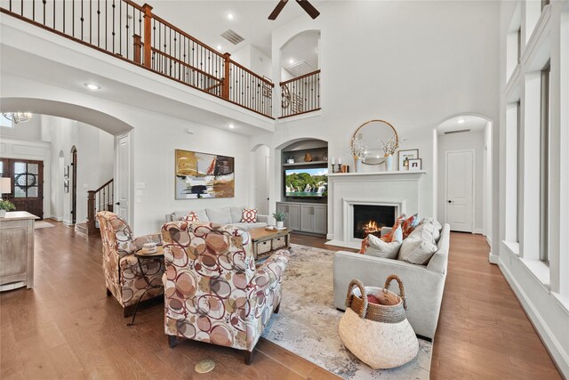 living room featuring visible vents, a towering ceiling, wood finished floors, a fireplace with flush hearth, and ceiling fan with notable chandelier