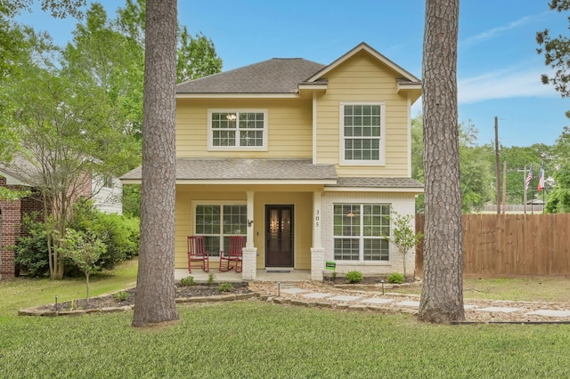 traditional-style house featuring a front lawn, roof with shingles, fence, and covered porch
