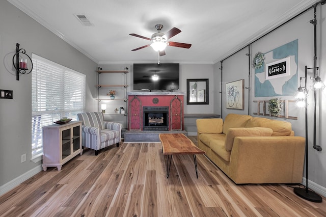 living area with visible vents, crown molding, wood finished floors, a fireplace with raised hearth, and a ceiling fan