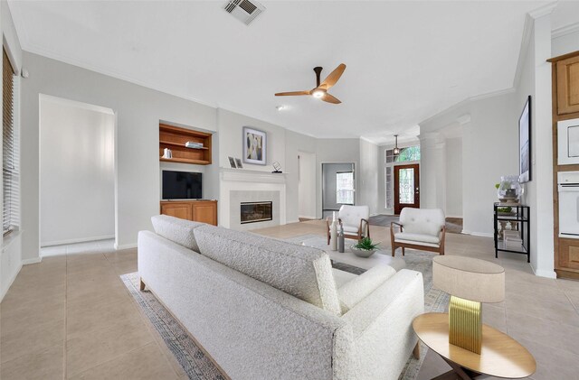 living area featuring crown molding, light tile patterned flooring, a glass covered fireplace, a ceiling fan, and visible vents