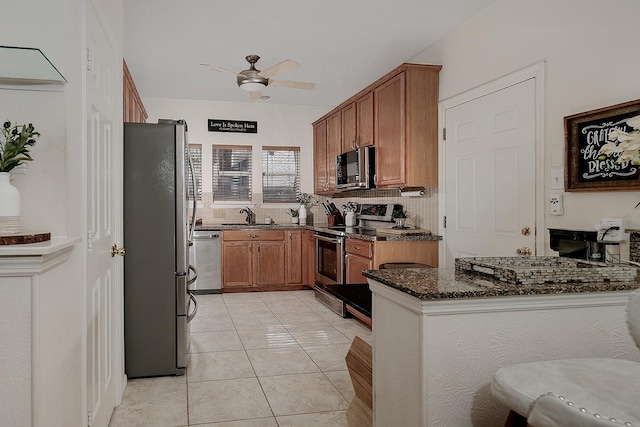 kitchen featuring ceiling fan, brown cabinets, a sink, dark stone countertops, and stainless steel appliances
