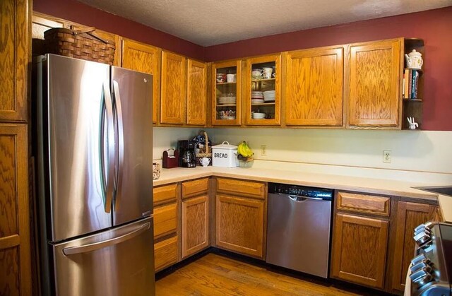 kitchen featuring brown cabinets, wood finished floors, light countertops, glass insert cabinets, and stainless steel appliances