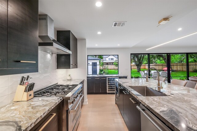 kitchen featuring wall chimney range hood, a sink, light wood finished floors, beverage cooler, and stainless steel appliances