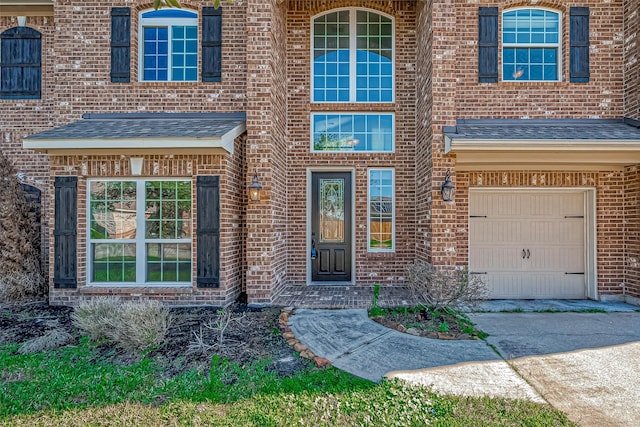 view of exterior entry with a garage, roof with shingles, and brick siding