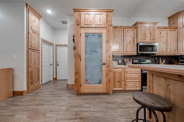 kitchen with backsplash, light wood-type flooring, light countertops, stainless steel microwave, and light brown cabinets