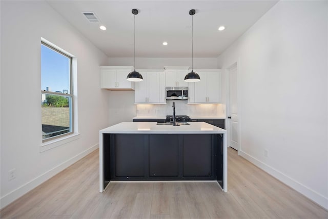 kitchen featuring decorative backsplash, a kitchen island with sink, stainless steel microwave, light wood finished floors, and white cabinetry