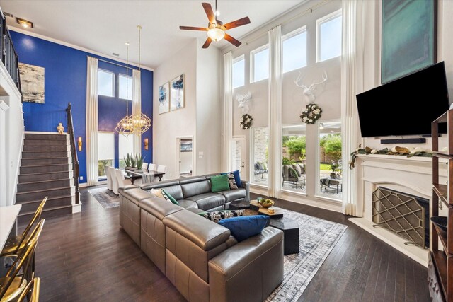 living area with stairway, a fireplace with raised hearth, ceiling fan with notable chandelier, dark wood-style floors, and a high ceiling