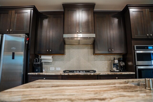 kitchen with dark brown cabinetry, under cabinet range hood, light stone counters, backsplash, and stainless steel appliances