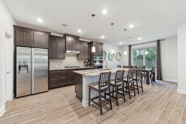 26 Heirloom Garden Pl, Magnolia, TX 77354 - kitchen featuring appliances with stainless steel finishes, dark brown cabinetry, a center island with sink, decorative backsplash, and light wood-style flooring
