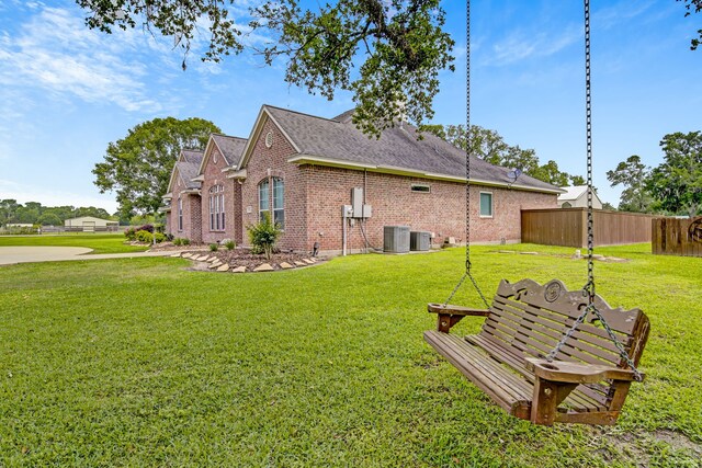 view of side of property with fence, roof with shingles, brick siding, central AC unit, and a lawn