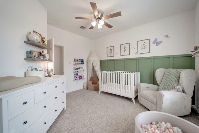 bedroom with ceiling fan, a crib, light carpet, a wainscoted wall, and visible vents