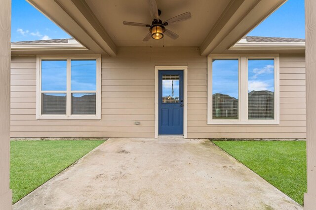 view of exterior entry with a lawn, a shingled roof, ceiling fan, and a patio area