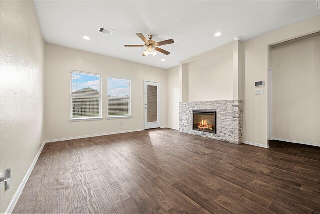 unfurnished living room with ceiling fan, a stone fireplace, baseboards, visible vents, and dark wood-style floors