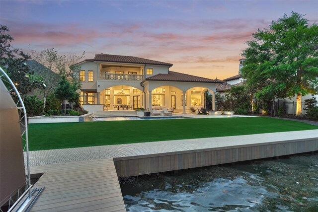 back of property at dusk featuring fence, a balcony, a patio, stucco siding, and a lawn