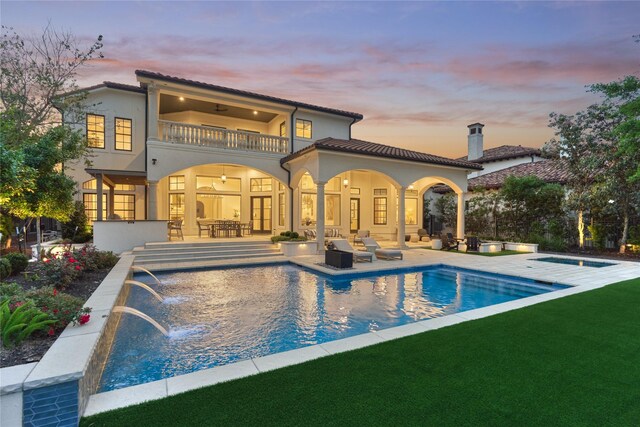 back of property at dusk with an outdoor pool, a patio area, a balcony, a yard, and stucco siding