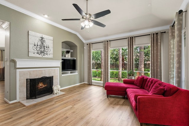 living room with ceiling fan, a tiled fireplace, crown molding, built in shelves, and wood finished floors