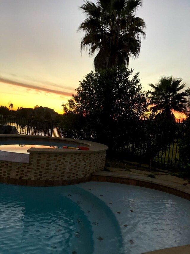 pool at dusk featuring fence and an outdoor hot tub