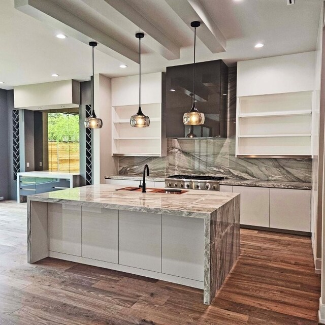 kitchen with open shelves, decorative backsplash, a center island with sink, dark wood-type flooring, and a sink