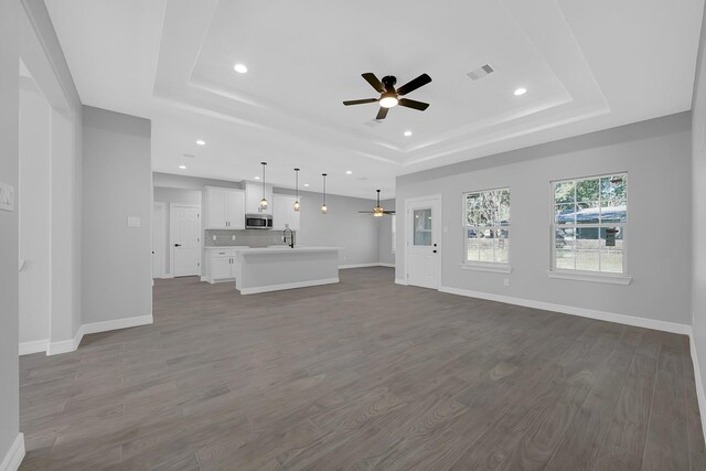 unfurnished living room featuring a ceiling fan, visible vents, a raised ceiling, baseboards, and dark wood-type flooring