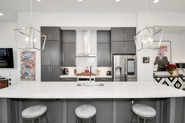 kitchen featuring stainless steel refrigerator with ice dispenser, wall chimney exhaust hood, a chandelier, gray cabinets, and backsplash