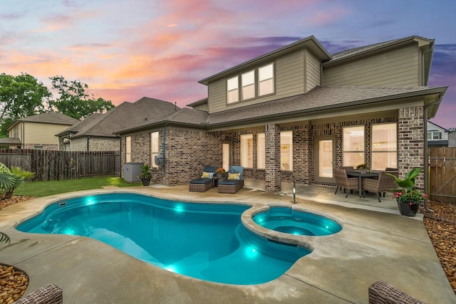 back of property at dusk featuring a pool with connected hot tub, a patio area, brick siding, a fenced backyard, and a shingled roof