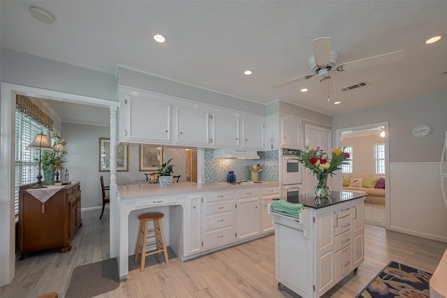 kitchen featuring light wood-type flooring, multiple ovens, under cabinet range hood, white cabinets, and a ceiling fan
