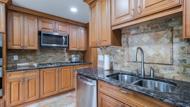 kitchen with brown cabinetry, a sink, dark stone countertops, appliances with stainless steel finishes, and backsplash