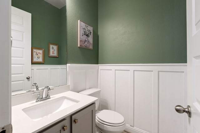 half bathroom with vanity, wainscoting, a decorative wall, and toilet