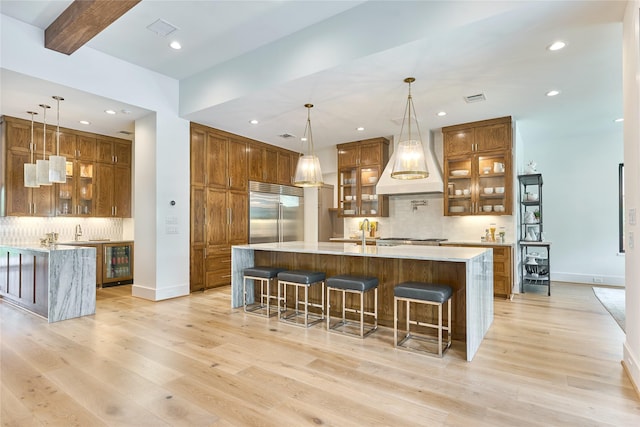 kitchen with a large island with sink, stainless steel built in fridge, custom exhaust hood, and brown cabinetry