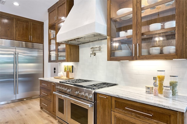 kitchen with light stone countertops, premium range hood, high quality appliances, and glass insert cabinets
