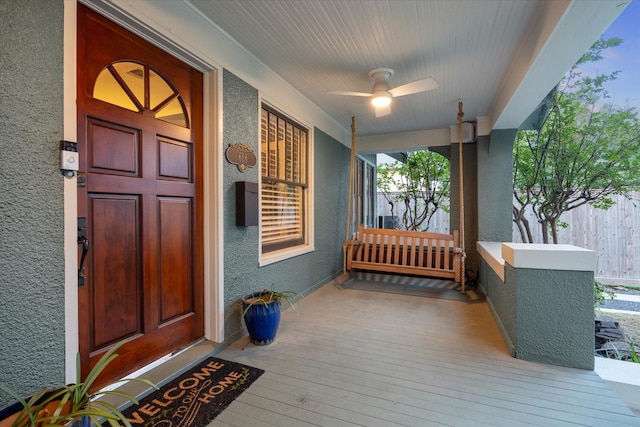 view of exterior entry featuring a porch, a ceiling fan, stucco siding, and fence