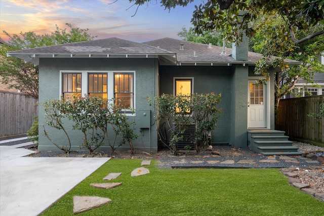 back of property with a lawn, a shingled roof, stucco siding, and fence