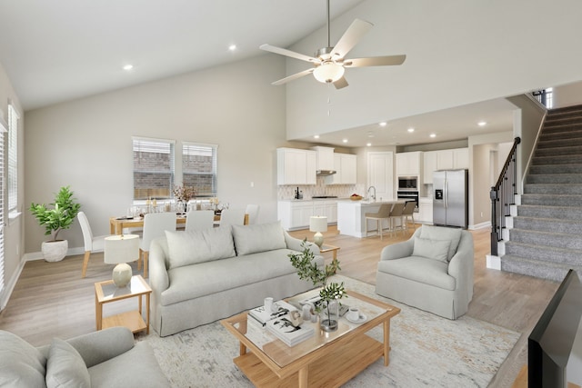 living area featuring light wood-style floors, baseboards, high vaulted ceiling, stairway, and a ceiling fan