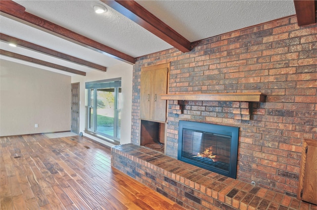 unfurnished living room with a brick fireplace, wood finished floors, beam ceiling, baseboards, and a textured ceiling