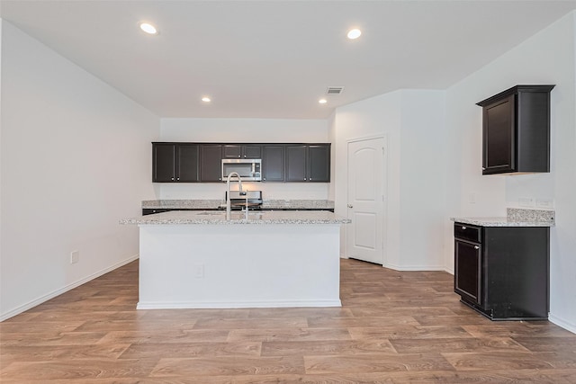 12518 Pelican Bay Dr, Houston, TX 77038 - kitchen featuring light stone countertops, a center island with sink, baseboards, stainless steel microwave, and light wood finished floors