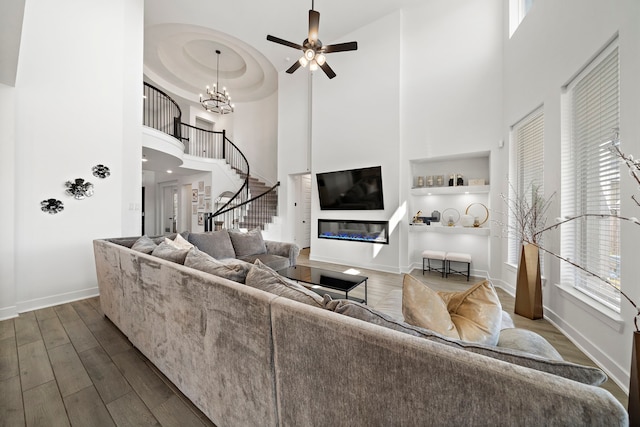 living room featuring a high ceiling, dark wood finished floors, a glass covered fireplace, ceiling fan with notable chandelier, and baseboards