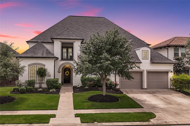 french provincial home with roof with shingles, an attached garage, stucco siding, concrete driveway, and a yard