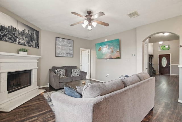 living room with ceiling fan, dark wood-style flooring, visible vents, arched walkways, and a fireplace with raised hearth