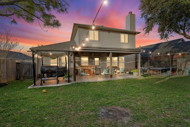 back of property at dusk featuring a trampoline, a fenced backyard, heating fuel, a chimney, and a patio