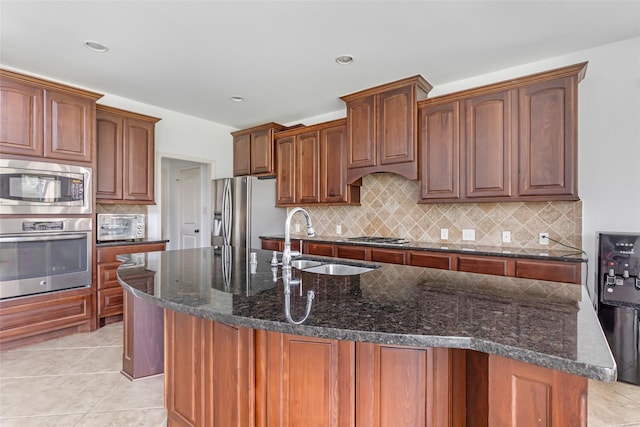 kitchen with a sink, dark stone counters, stainless steel appliances, tasteful backsplash, and a center island with sink