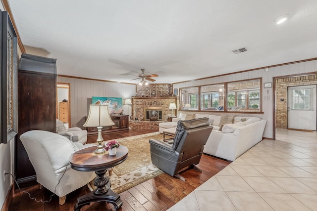 tiled living area with crown molding, ceiling fan, a fireplace, wood walls, and visible vents