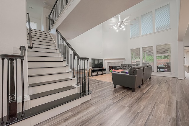 living area featuring a high ceiling, a glass covered fireplace, stairs, a ceiling fan, and wood finished floors