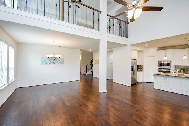 unfurnished living room featuring baseboards, ceiling fan with notable chandelier, dark wood-style floors, a high ceiling, and stairs