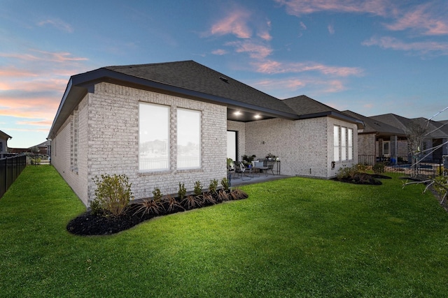 back of property at dusk featuring fence, roof with shingles, brick siding, a patio area, and a lawn