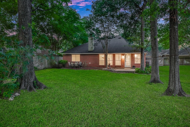 back of house at dusk with a lawn, brick siding, fence, and a chimney