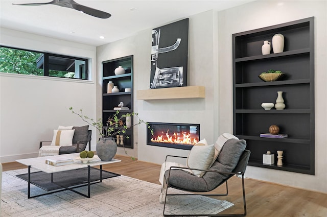living room with ceiling fan, a glass covered fireplace, built in features, and wood finished floors