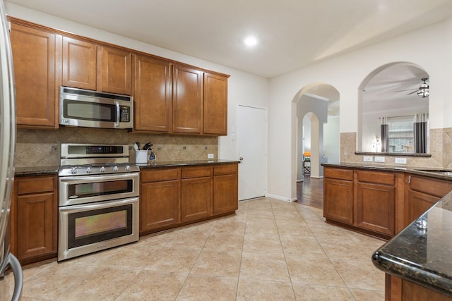 kitchen featuring appliances with stainless steel finishes, arched walkways, brown cabinetry, dark stone counters, and decorative backsplash